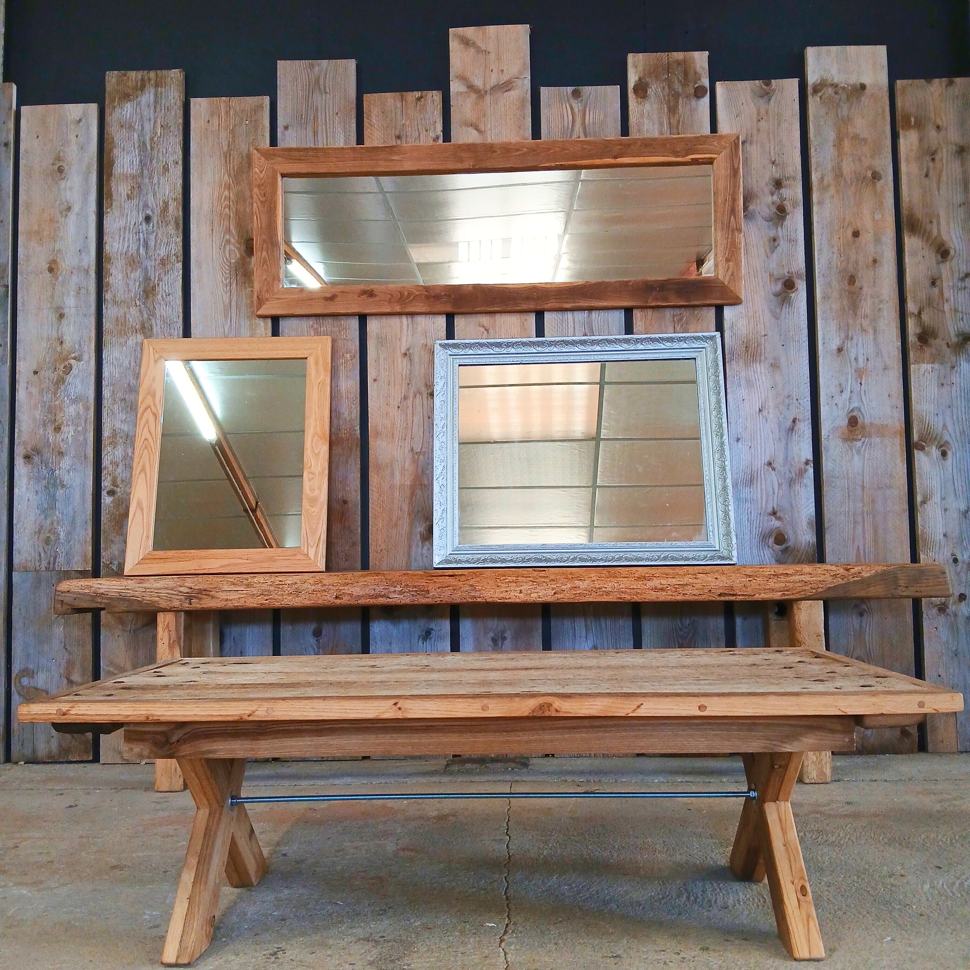 Rustic reclaimed wood mirror collection staged against a black accent wall with vertical scaffolding planks. The set includes two solid oak mirrors and one painted silver-framed mirror of unknown wood. Displayed alongside the ‘Charente’ reclaimed oak console table, originally a carpenter’s workbench with teak bow tie joins, and the ‘Lisonne’ coffee table crafted from a reclaimed solid oak door.
